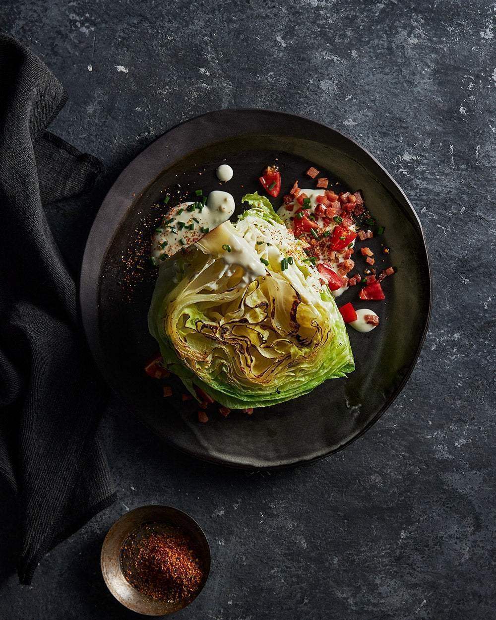 Grilled wedge salad with dressing, diced tomatoes, bacon, and chives on dark plate by Marion Grasby