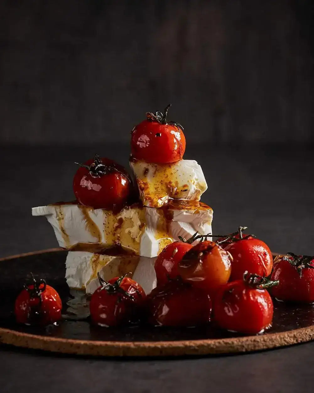 Roasted cherry tomatoes on creamy feta cheese with oil drizzle on dark plate, Marion Grasby recipe