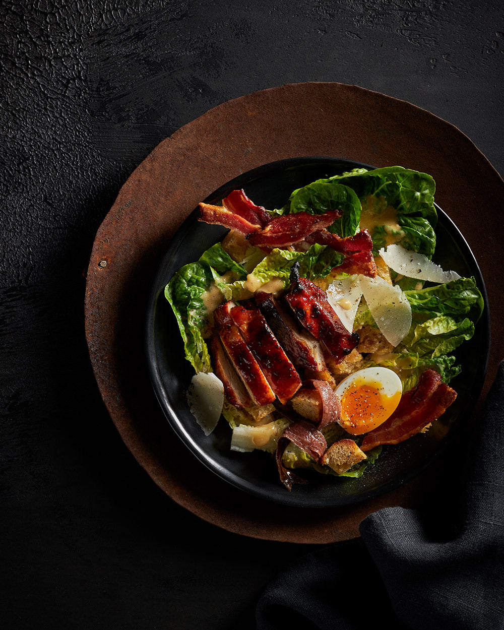 Caesar salad with grilled chicken, crispy bacon, soft-boiled egg, parmesan, and croutons on dark plate