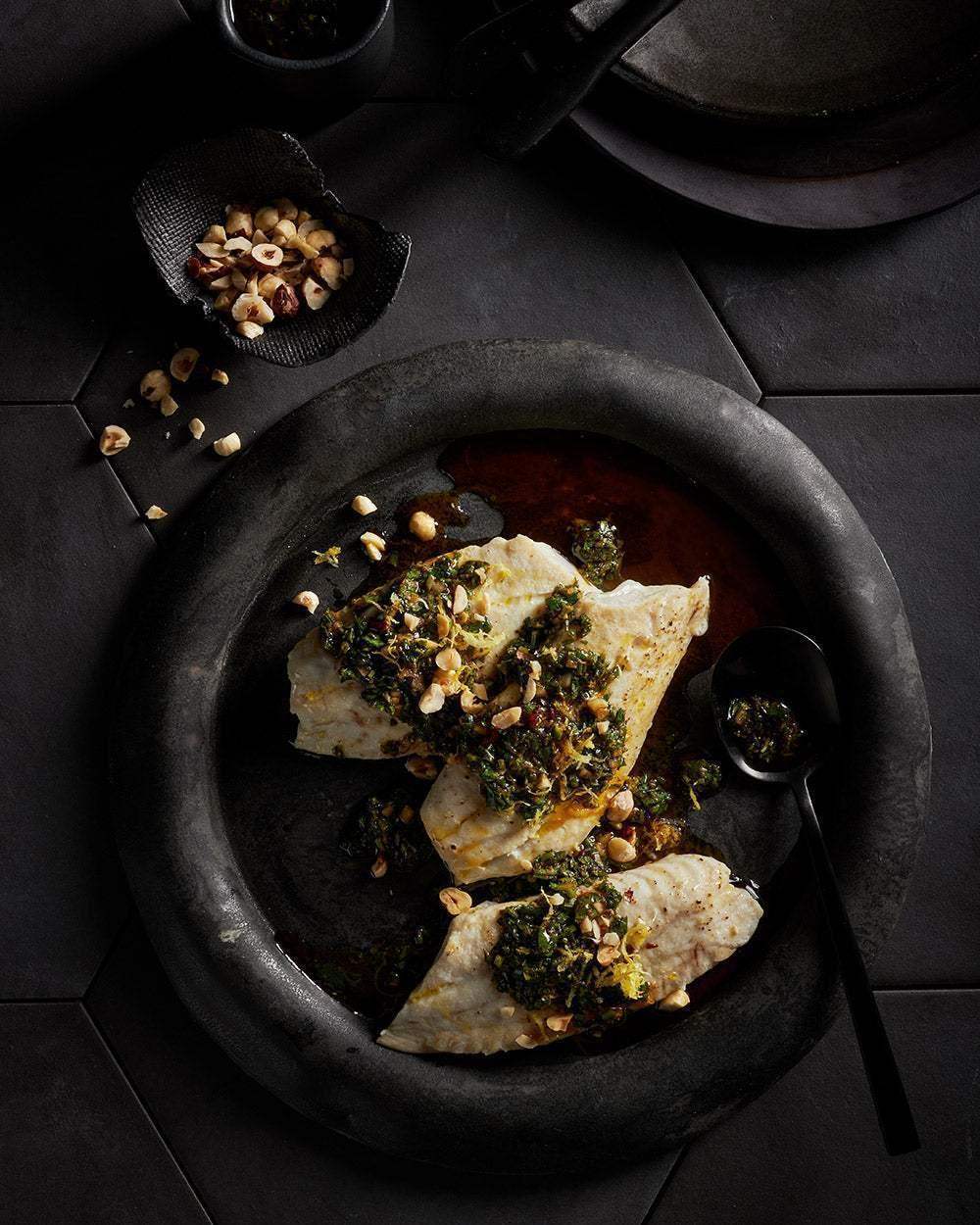 Cooked white fish fillets with herb sauce and chopped nuts on a dark plate, styled by Marion Grasby