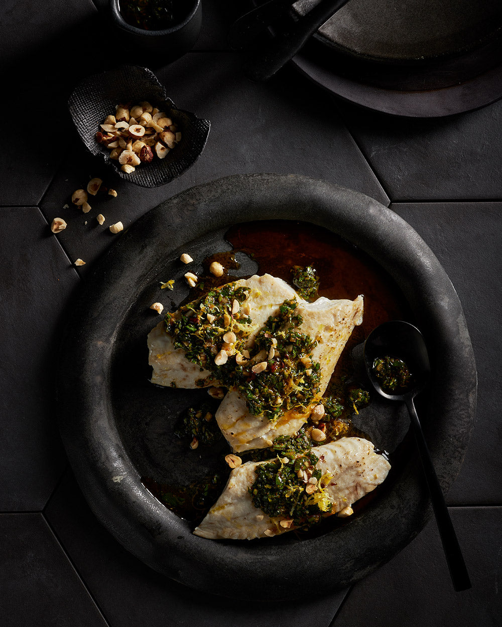 Grilled white fish fillets topped with herb sauce and chopped nuts on a dark plate