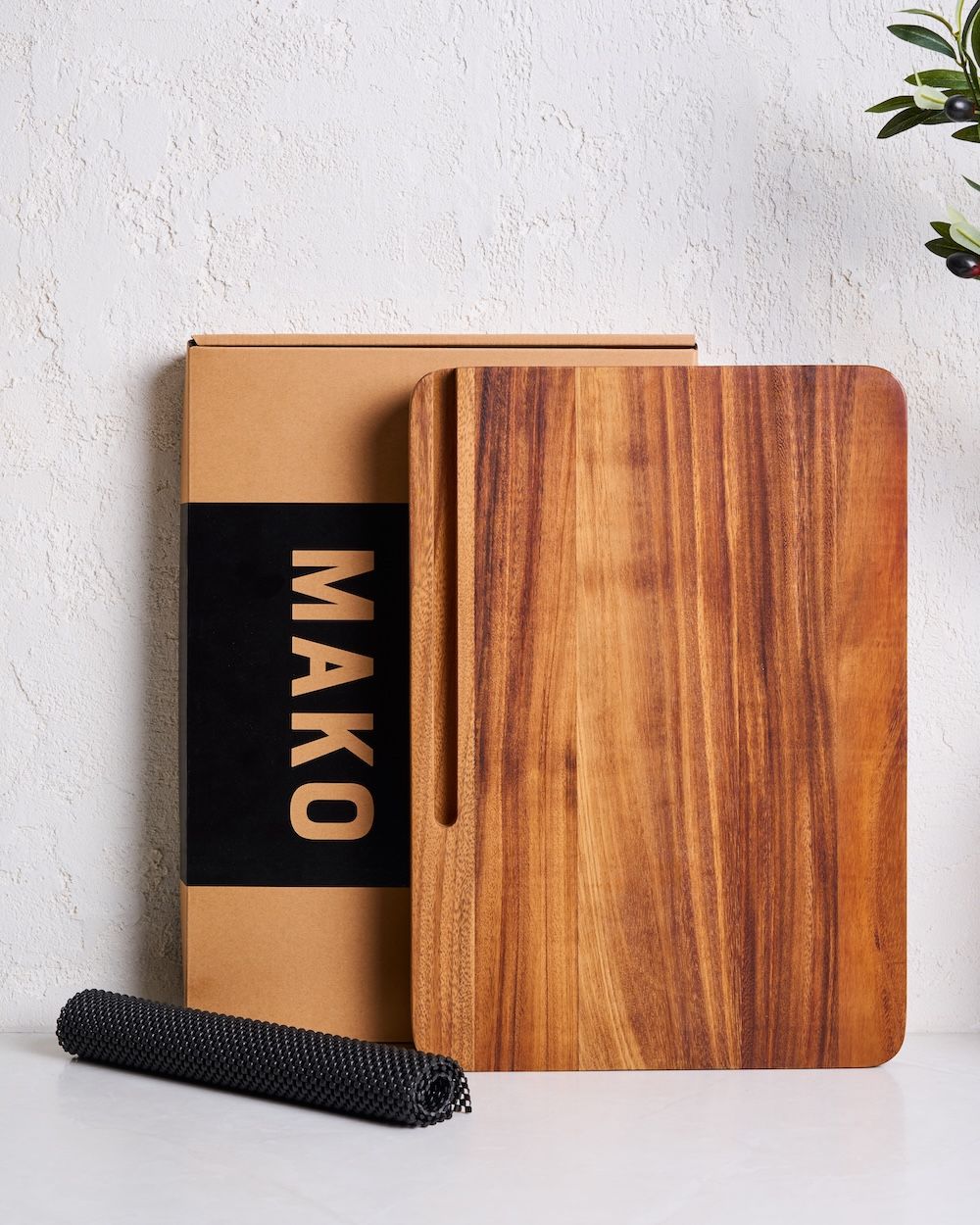 Acacia wood cutting board with juice groove, black non-slip mat, and box on white counter