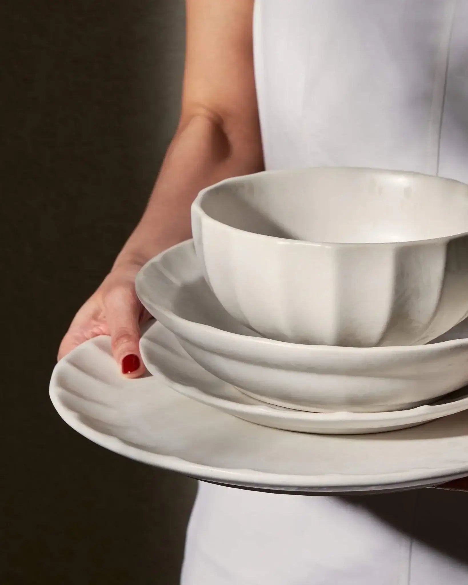 Hand holding stacked white ceramic bowl and plates with scalloped edges against dark background
