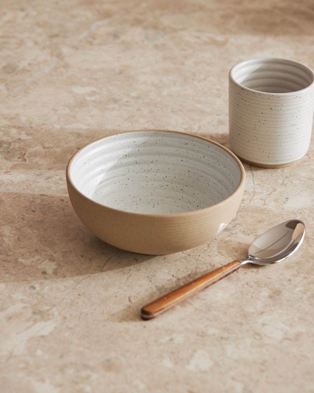 Speckled ceramic cereal bowl and cup with wooden-handled spoon on stone countertop