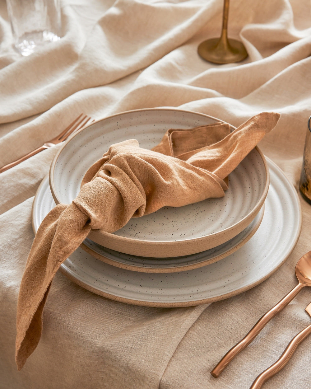 Set of four beige stoneware wide bowls stacked on a neutral linen tablecloth with gold cutlery and a tan cloth napkin.