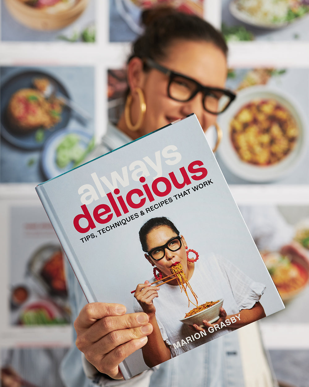 Person holding 'Always Delicious' cookbook with noodle dish cover and food photos in background.