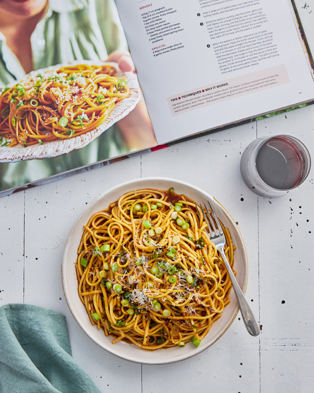 Open cookbook with a plate of saucy noodles topped with scallions and grated cheese, beside a glass of red wine.