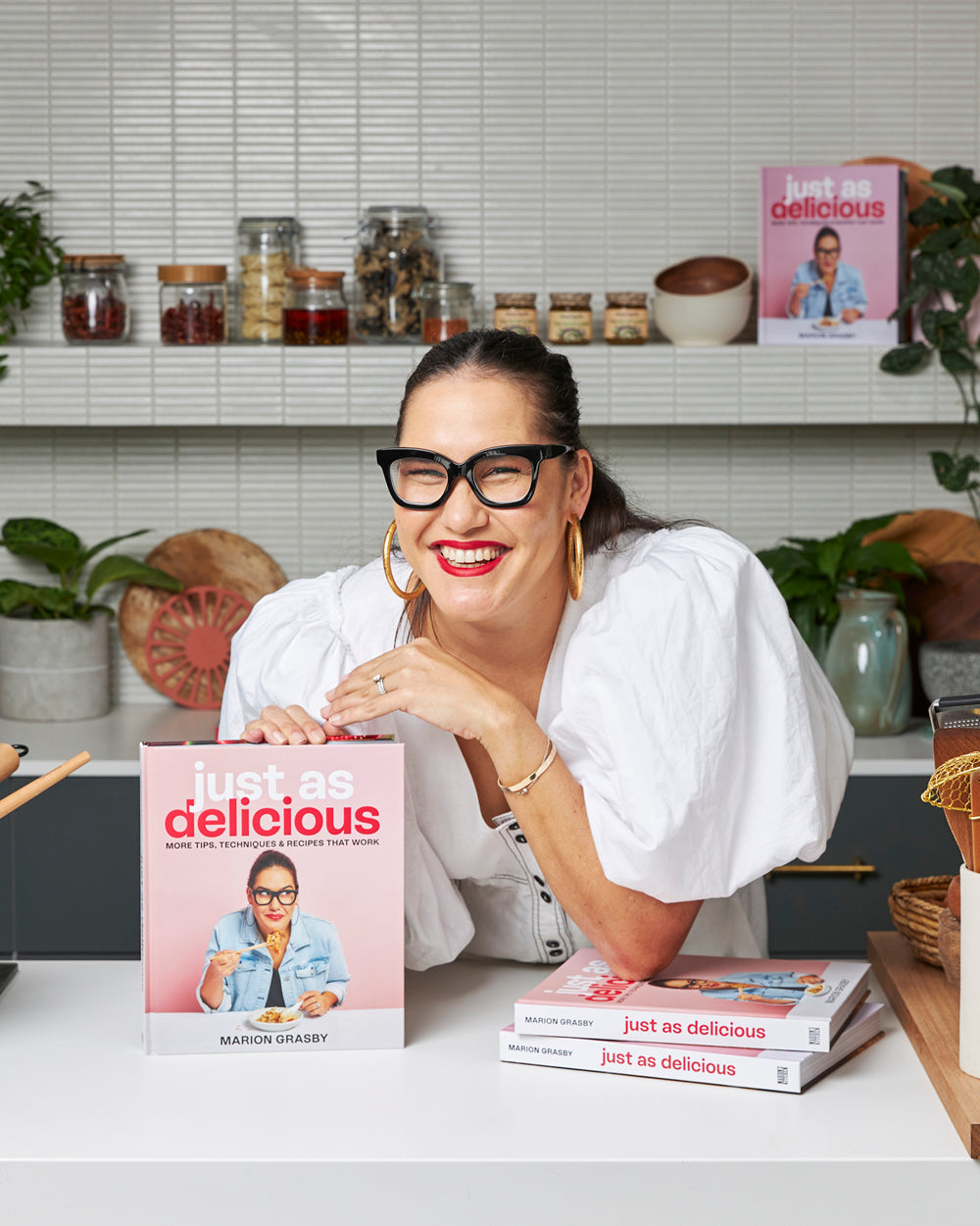 Smiling woman with glasses holding 'Just As Delicious' cookbook in modern kitchen setting.