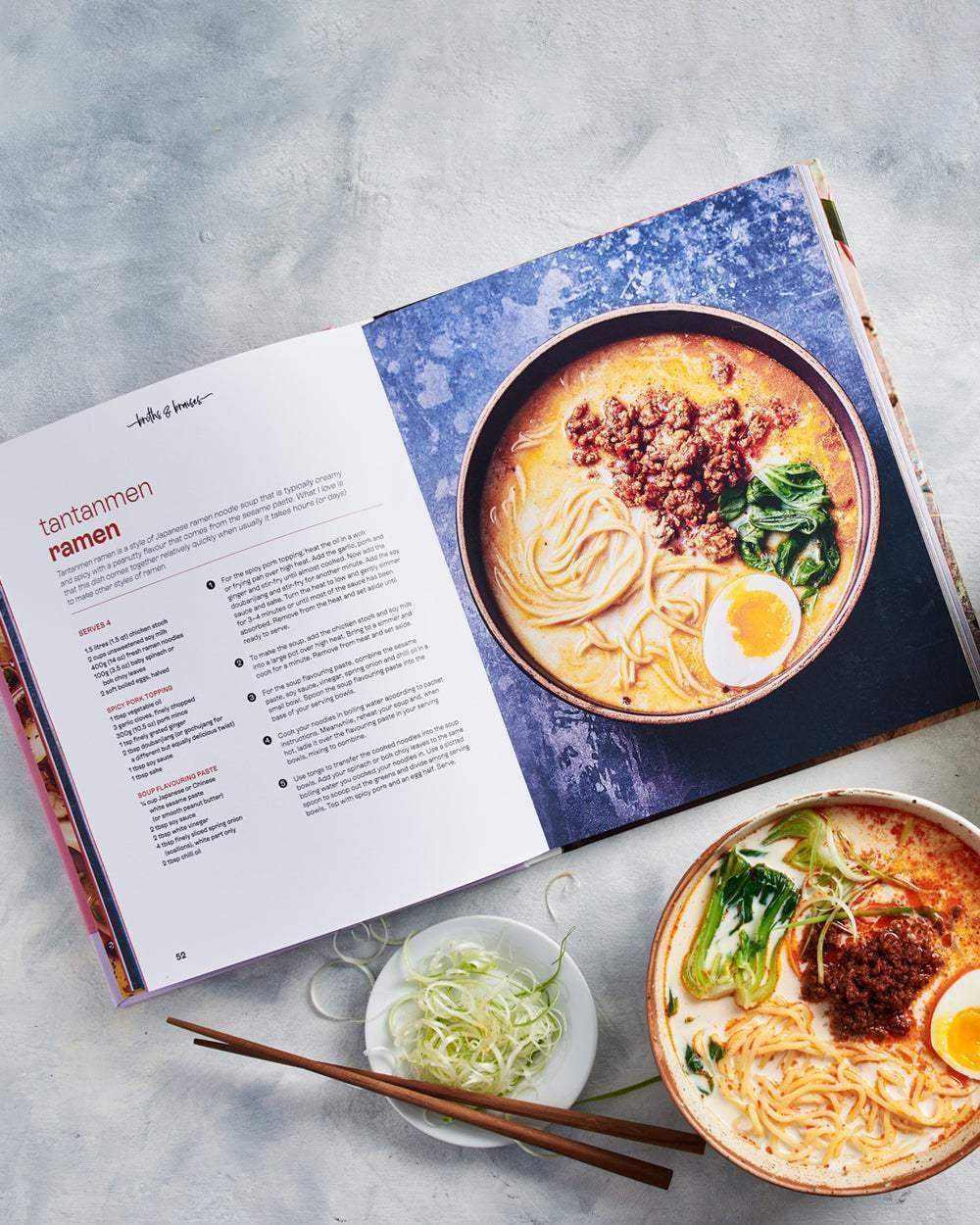 Open cookbook by Marion Grasby showing tantanmen ramen recipe with a bowl of spicy ramen and chopsticks on a light surface