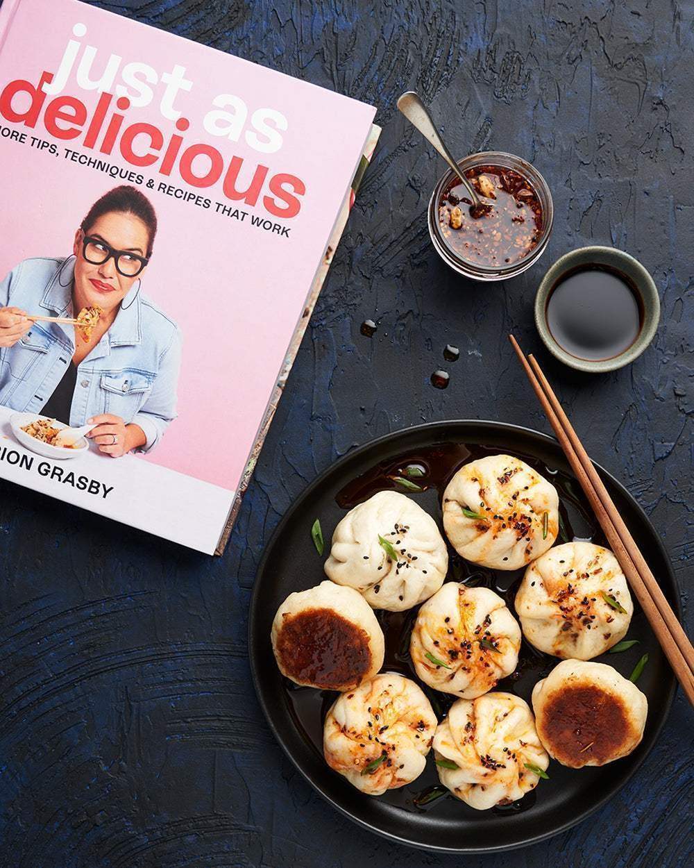 Marion Grasby's cookbook Just As Delicious next to a black plate of pan-fried dumplings with dipping sauce and chopsticks
