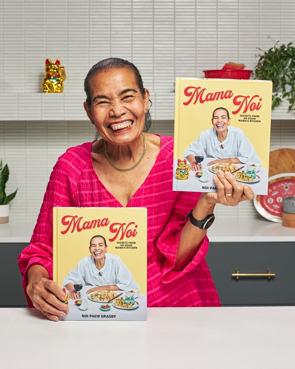 Smiling woman in pink top holding two 'Mama Noi' Asian cookbook covers in a modern kitchen