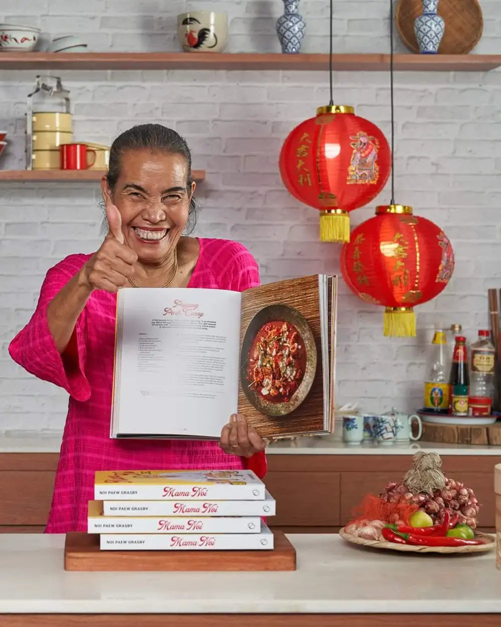 Woman in pink shirt shows open Asian cookbook with spicy pork curry recipe, red lanterns and ingredients