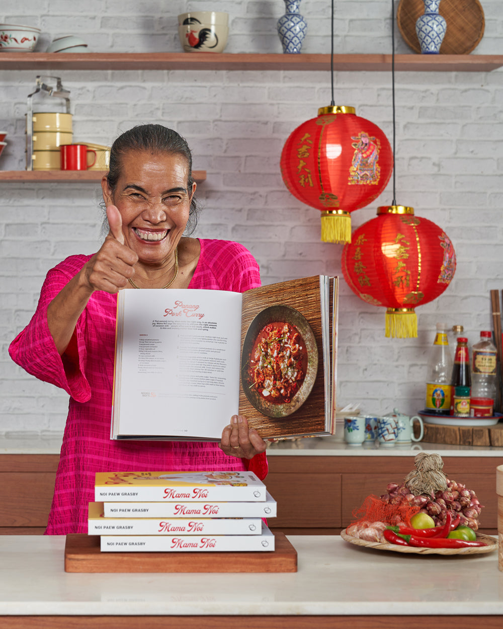 Smiling woman in pink holding open Asian cookbook in kitchen with red lanterns and spices.