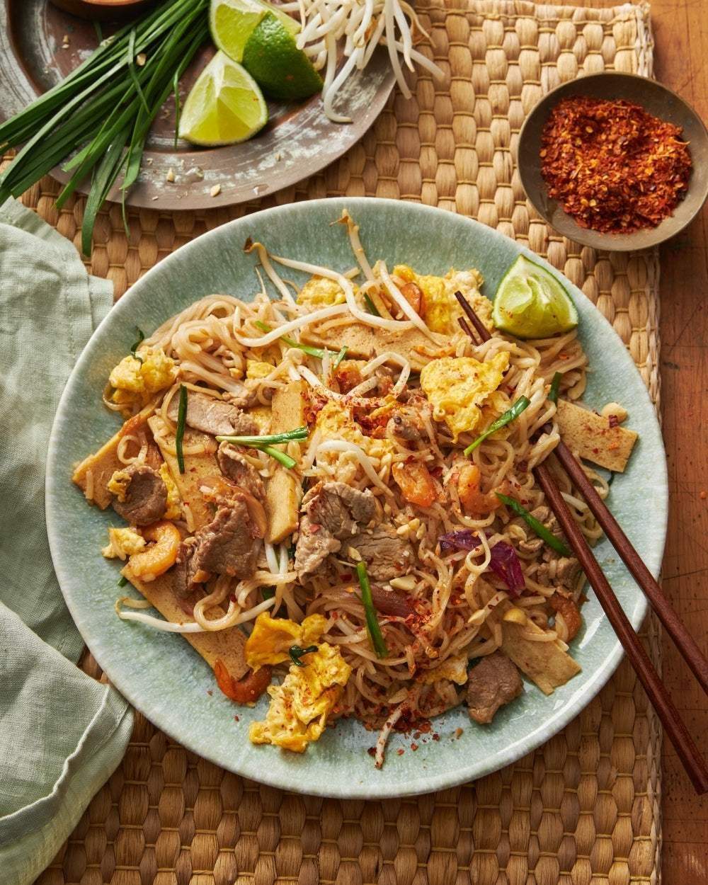 Plate of Asian stir-fried noodles with beef, shrimp, egg, bean sprouts, and lime wedges, chopsticks on woven mat