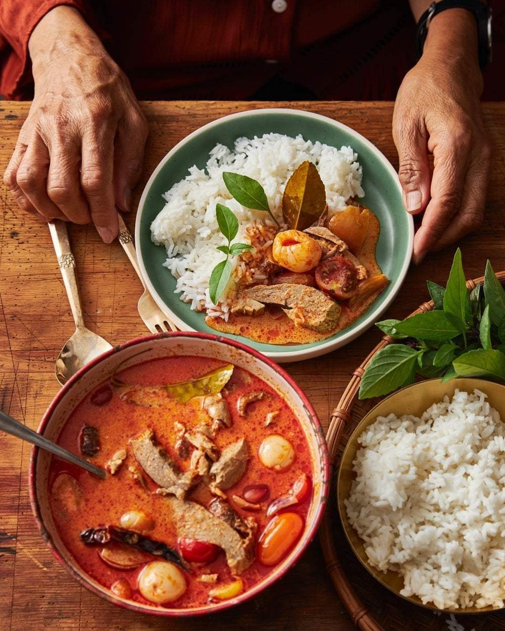 Hands holding a green plate with rice and Asian red curry, served with a bowl of curry and rice on a wooden table
