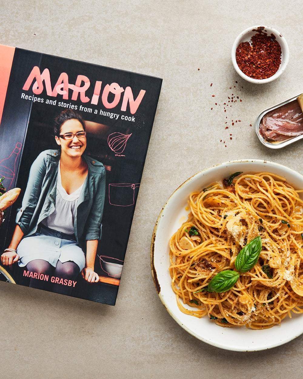 Marion Grasby's cookbook 'Marion Recipes and stories from a hungry cook' beside a plate of spaghetti with basil and a bowl of chili flakes