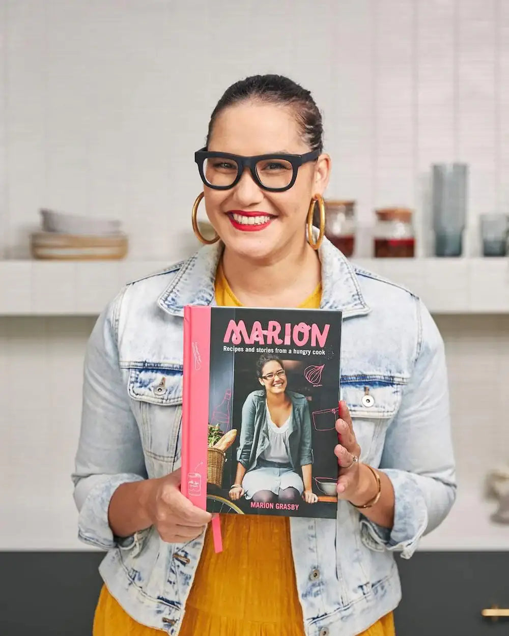 Marion Grasby holding her cookbook Marion with kitchen background, wearing glasses and denim jacket