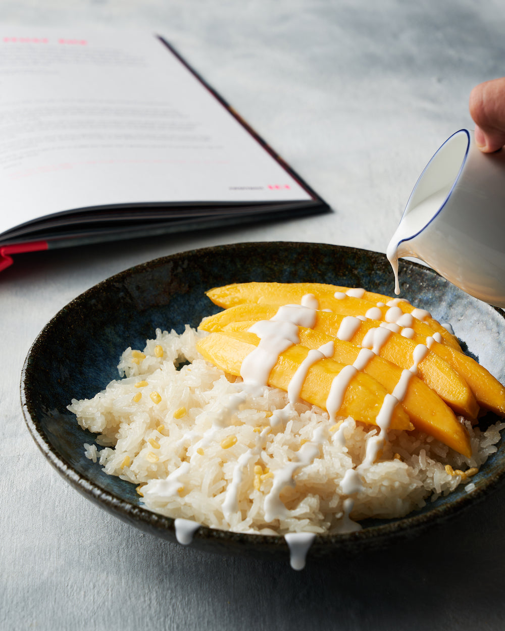Mango sticky rice dessert with coconut sauce and an open cookbook in the background.